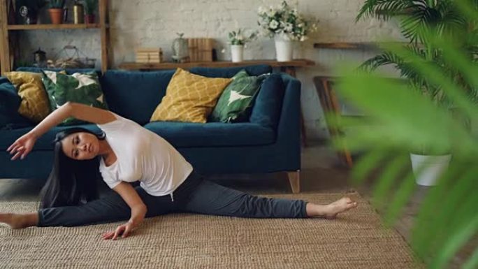 灵活的亚洲女孩坐在地板上在家做瑜伽，伸展腿和身体独自锻炼。带家具和植物的美丽房间是背景。
