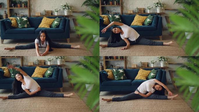 灵活的亚洲女孩坐在地板上在家做瑜伽，伸展腿和身体独自锻炼。带家具和植物的美丽房间是背景。