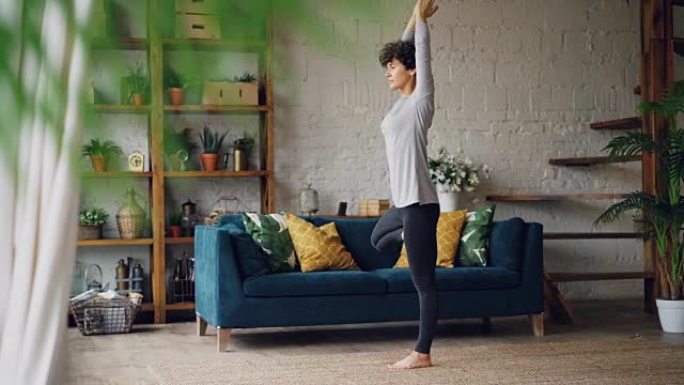 灵活而强壮的年轻女子正在家里训练瑜伽，站在一条腿上，保持双臂的平衡。健康的生活方式和室内设计理念。