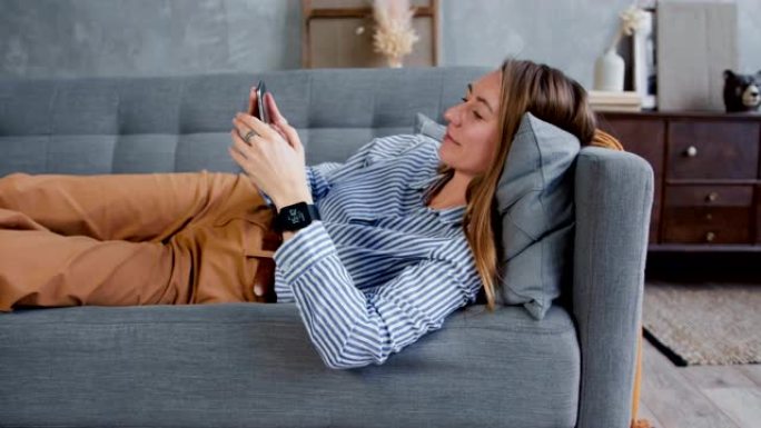 智能手机成瘾。年轻美丽微笑的金发女人在家里使用社交网络应用程序在沙发上拖延。