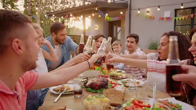 家人和朋友聚集在餐桌旁，举起玻璃杯和瓶子敬酒并碰杯。大家庭花园派对庆典。