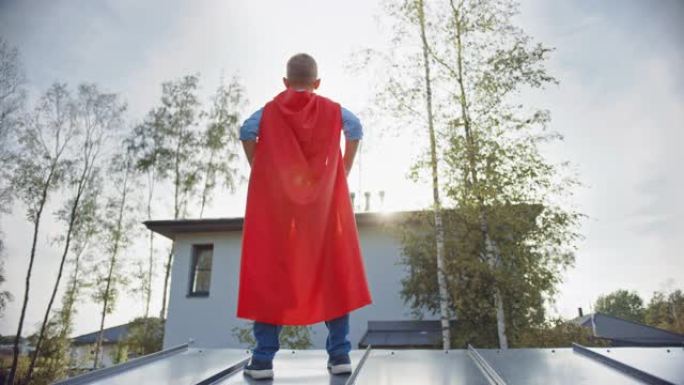 男孩正在扮演一个超级英雄的角色。他站在房子的屋顶上，双手放在腰上。年轻人穿着鲜红色的斗篷。他在看太阳