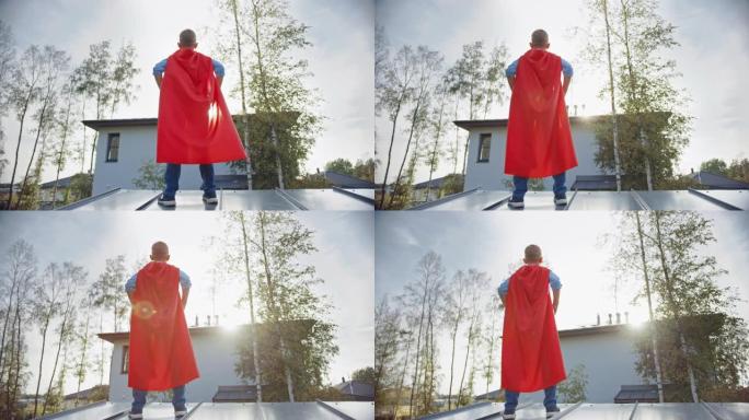 男孩正在扮演一个超级英雄的角色。他站在房子的屋顶上，双手放在腰上。年轻人穿着鲜红色的斗篷。他在看太阳