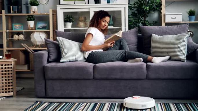 漂亮的非洲裔美国学生正在打开机器人胡佛，一边看书，一边享受业余爱好，一边在沙发上放松，一边清洁地板。