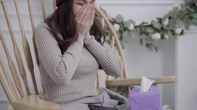 亚洲妇女感到头痛裹在灰色毯子在家里的客厅放松时，躺在椅子上吹鼻子和使用纸巾。圣诞节和新年假期概念中的