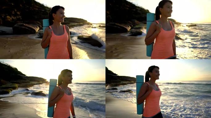 宁静的拉丁美洲妇女在沙滩上散步，同时拿着瑜伽垫微笑着