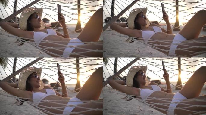 年轻的亚洲妇女在日落时海边的吊床上看手机