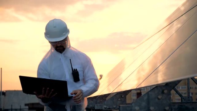 环保能源概念。男性太阳能建筑商站在太阳能电池板附近时正在操作笔记本电脑。