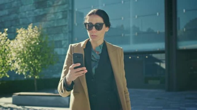 优雅的女商人在现代玻璃建筑前行走时使用智能手机开展业务。穿着外套和墨镜的美丽时尚女性在现代城市环境中