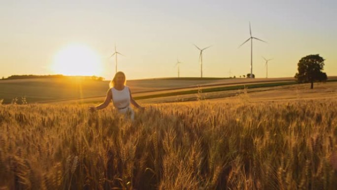 快乐的女人一边抚摸着小麦，一边跑在远处有风力涡轮机的田地里
