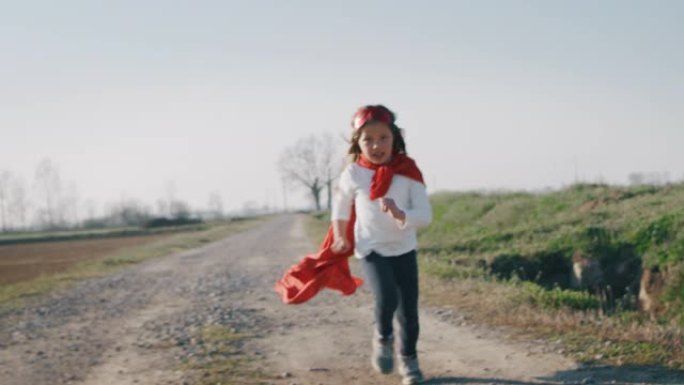 一个有趣的小女孩打扮成超级英雄的真实照片，穿着红色斗篷跑步，在阳光明媚的日子里玩得开心