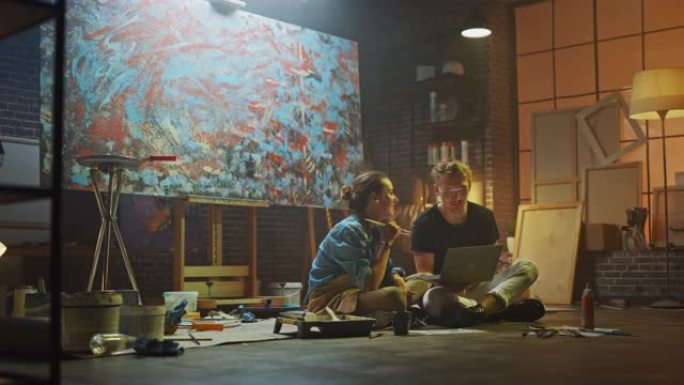 坐在工作室里的几位艺术家使用笔记本电脑，从事一个项目，交谈和微笑。画家和她的搭档做研究。带油画和工具