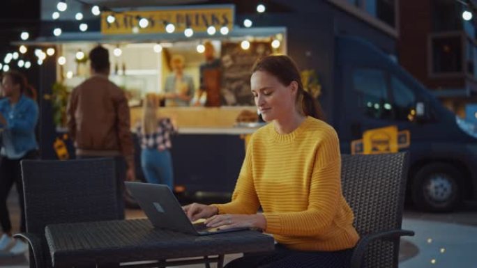 美丽的黑发年轻女子坐在户外街头美食咖啡馆的桌子旁，正在笔记本电脑上工作。她正在浏览互联网或社交媒体，