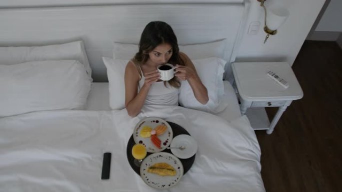 美丽的女人在酒店看电视时在床上享受早餐