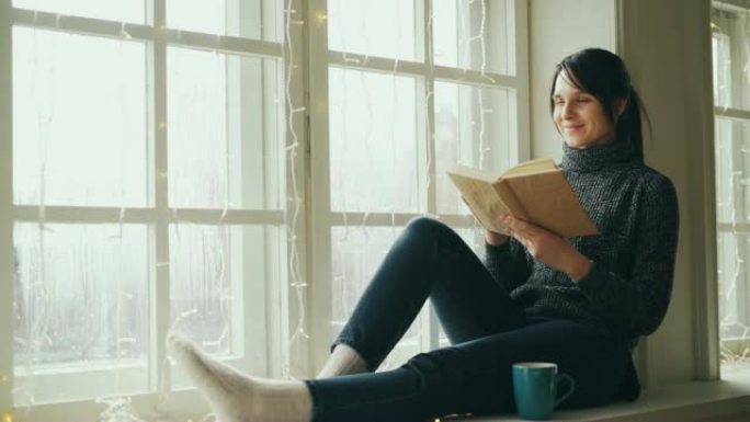 梦幻般的年轻女子正在看书，圣诞节时坐在窗台上微笑着，窗台上装饰着照明。女孩穿着毛衣和时髦的牛仔裤。
