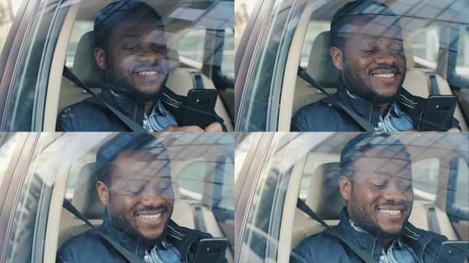 英俊的黑人坐在汽车上，坐在乘客座位上，用一只手使用智能手机，输入消息，通过互联网浏览。从车外拍摄的摄