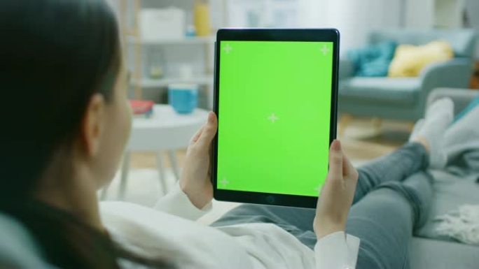 年轻女子在家休息在沙发上使用绿色模拟屏幕平板电脑在垂直肖像模式。女人使用平板电脑设备，浏览互联网，观