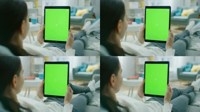 年轻女子在家休息在沙发上使用绿色模拟屏幕平板电脑在垂直肖像模式。女人使用平板电脑设备，浏览互联网，观