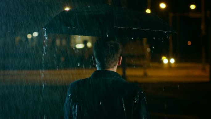 一个年轻的高加索短毛男人的照片，穿着牛仔裤外套在雨中撑着雨伞行走。他晚上在城里散步。