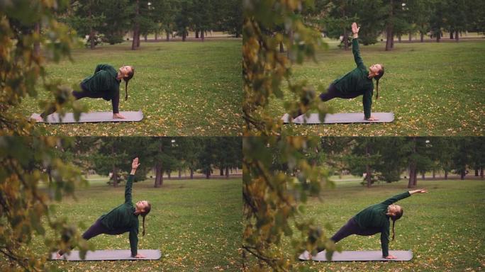 瑜伽学生正在公园瑜伽垫上练习侧角位置的变化。可以看到绿色和黄色的树木，叶子和草。