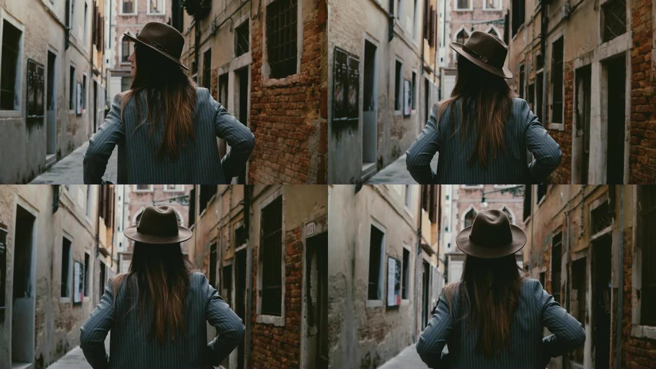 穿着帽子和西装的自信时尚年轻女商人沿着意大利威尼斯狭窄的古街行走的后景。
