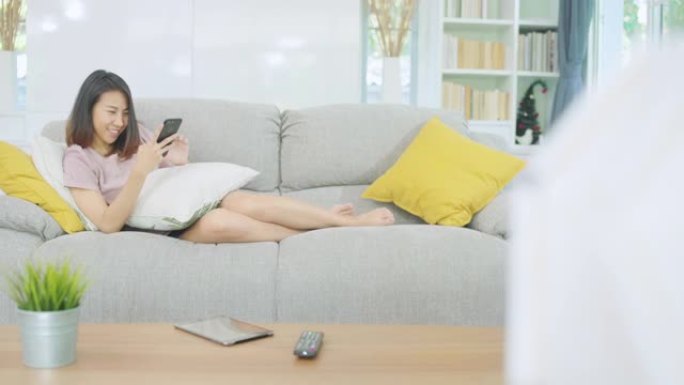 年轻的亚洲妇女使用智能手机检查社交媒体感到快乐的微笑，而躺在沙发上放松时，在家里的客厅。生活方式拉丁