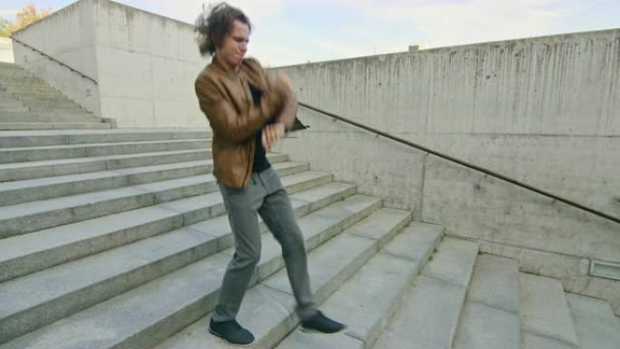 开朗快乐的长发年轻人在走下楼梯时积极跳舞。他穿着一件棕色皮夹克。在商务中心旁边的城市混凝土公园拍摄的