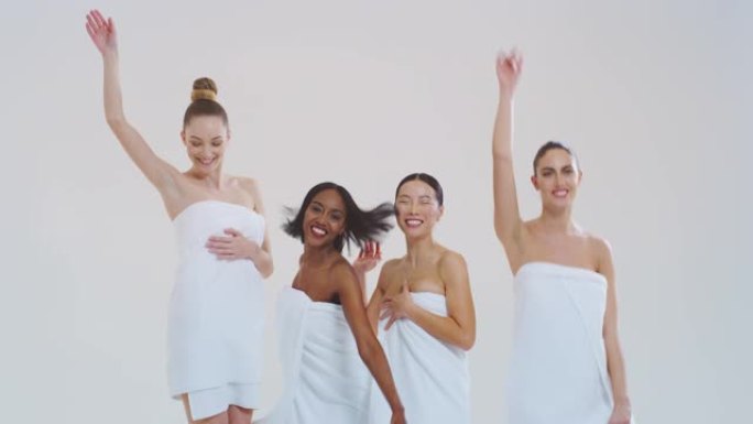 不同种族的美丽年轻微笑女性的慢动作，完美结实苗条的身体，穿着白色浴巾跳舞，享受孤立在白色背景上的乐趣