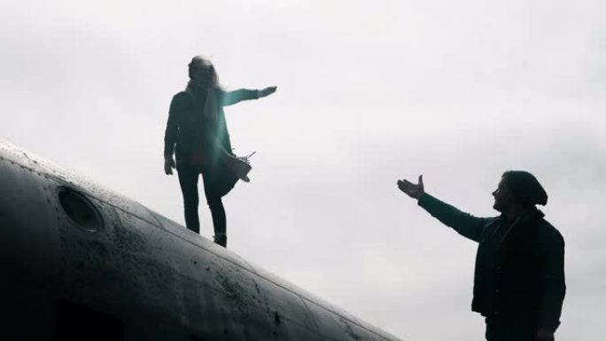 年轻女子站在冰岛坠毁的DC-3飞机的顶部，伸出手向站在附近的男子伸出手。