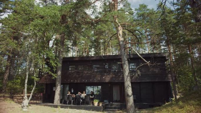 欧洲松树林中一座两层黑色木制小屋的风景照片。年轻的朋友们聚集在露台上。这是一个温暖阳光明媚的夏日早晨