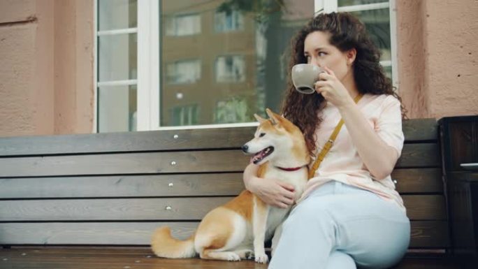 迷人的女孩在咖啡馆户外喝茶，坐在长凳上拍狗