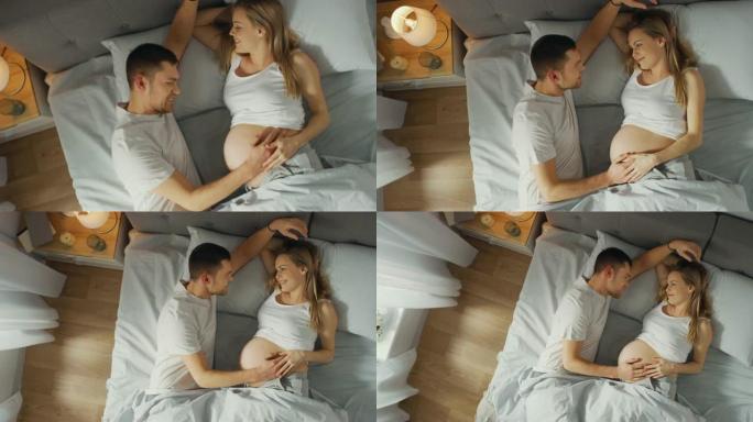 幸福的年轻夫妇在床上拥抱在一起，年轻的女人怀孕了，有爱心的伴侣温柔地抚摸着她的肚子。俯视图缩小相机拍