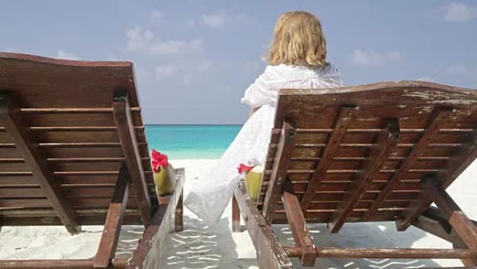 马尔代夫阳光明媚、宁静的热带海洋海滩，女士在躺椅上喝椰子饮料放松