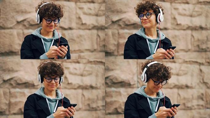 漂亮的女孩是耳机和眼镜正在听音乐和触摸智能手机屏幕选择歌曲站在城市的户外，背景是石墙。