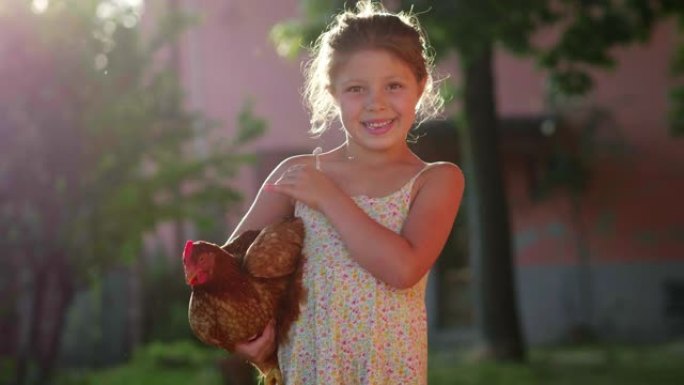 快乐的微笑小女孩的真实时刻在乡村房屋外抱着一只棕色的母鸡