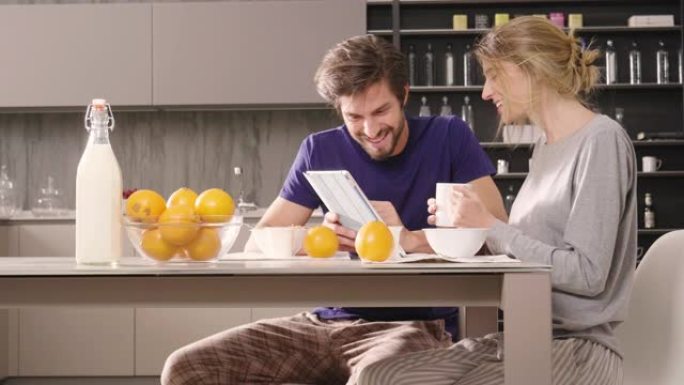厨房里的一对夫妇一边吃早餐，一边开心地微笑着看着平板电脑，一边看着他们假期或过去的纪念品照片。