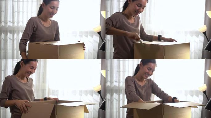 漂亮的年轻亚洲女人在家站在明亮的客厅的桌子上打开纸箱，在线送货