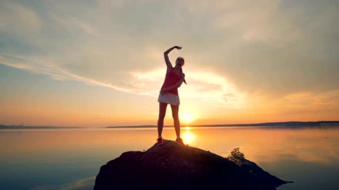 女运动员在湖边的一块岩石上热身。一个女孩在日落背景下跑步前伸展身体。
