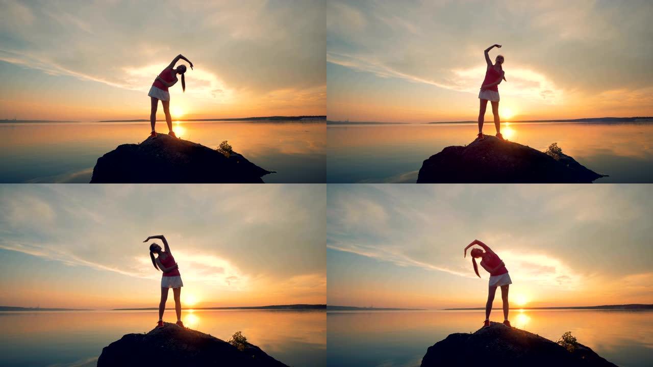 女运动员在湖边的一块岩石上热身。一个女孩在日落背景下跑步前伸展身体。