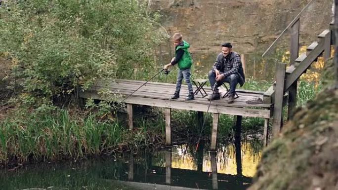严肃的孩子站在木墩和钓鱼竿上，而他慈爱的父亲坐在椅子上，抓鱼，微笑着看着儿子。