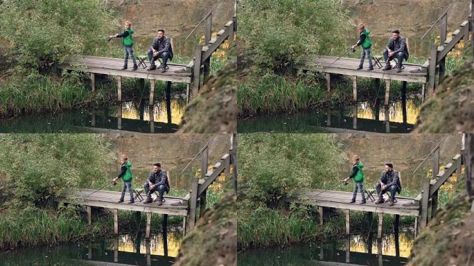 严肃的孩子站在木墩和钓鱼竿上，而他慈爱的父亲坐在椅子上，抓鱼，微笑着看着儿子。