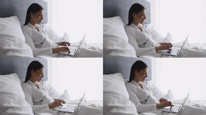快乐迷人的女士早上坐在床上使用笔记本电脑