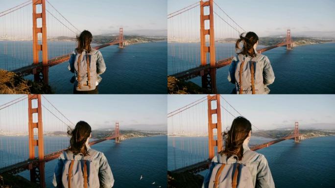后视图相机放大了背着背包的年轻女子，在金门大桥的风中吹着头发享受日落。
