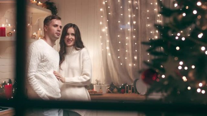 一对年轻夫妇在温暖舒适的家中看着圣诞树。他们期待这个圣诞节晚上的客人。摄像机穿过房子外面的窗户。