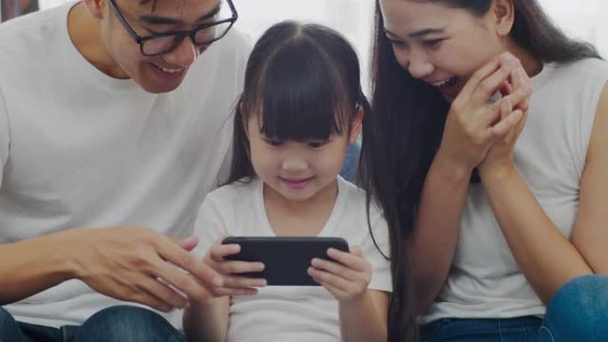 慢动作特写镜头: 幸福温暖的家庭父亲，母亲和女儿在智能手机上在线玩有趣的游戏，一起坐在沙发上，父母带