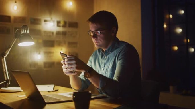专业人士晚上坐在办公室工作室的办公桌前，在笔记本电脑上工作。男子使用智能手机与数据，分析统计数据。3