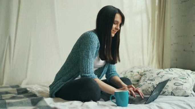 开朗的女学生正在家里学习，计算机坐在床上看着屏幕，打字和微笑。在线教育和房屋概念。