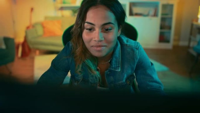 美丽而兴奋的黑人玩家女孩正在她的个人电脑上玩在线视频游戏。房间和电脑有彩色温暖的霓虹灯。在家舒适的夜
