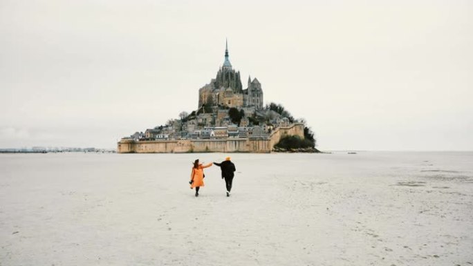无人机摄像机跟随年轻幸福的夫妇手牵着手奔向法国史诗般的圣米歇尔要塞岛。