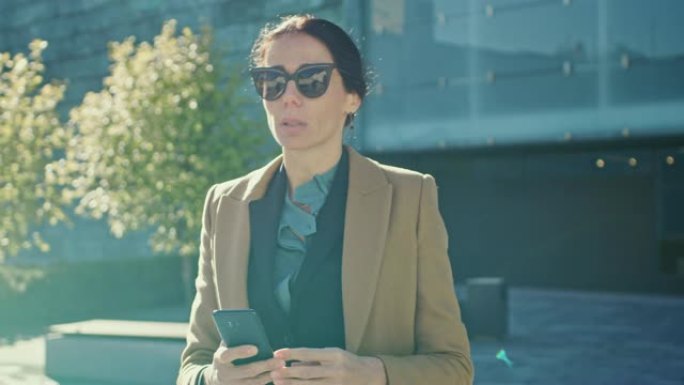 优雅的女商人在现代玻璃建筑前行走时使用智能手机开展业务。穿着外套和墨镜的美丽时尚女性在现代城市环境中